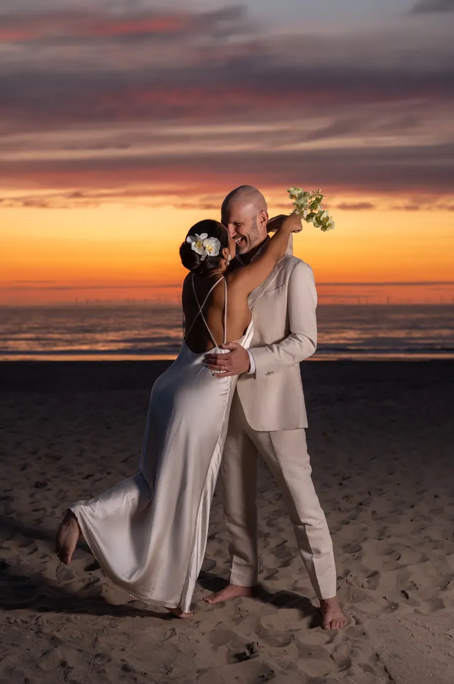 Casal abraçado à beira-mar, com o horizonte alaranjado do pôr do sol ao fundo