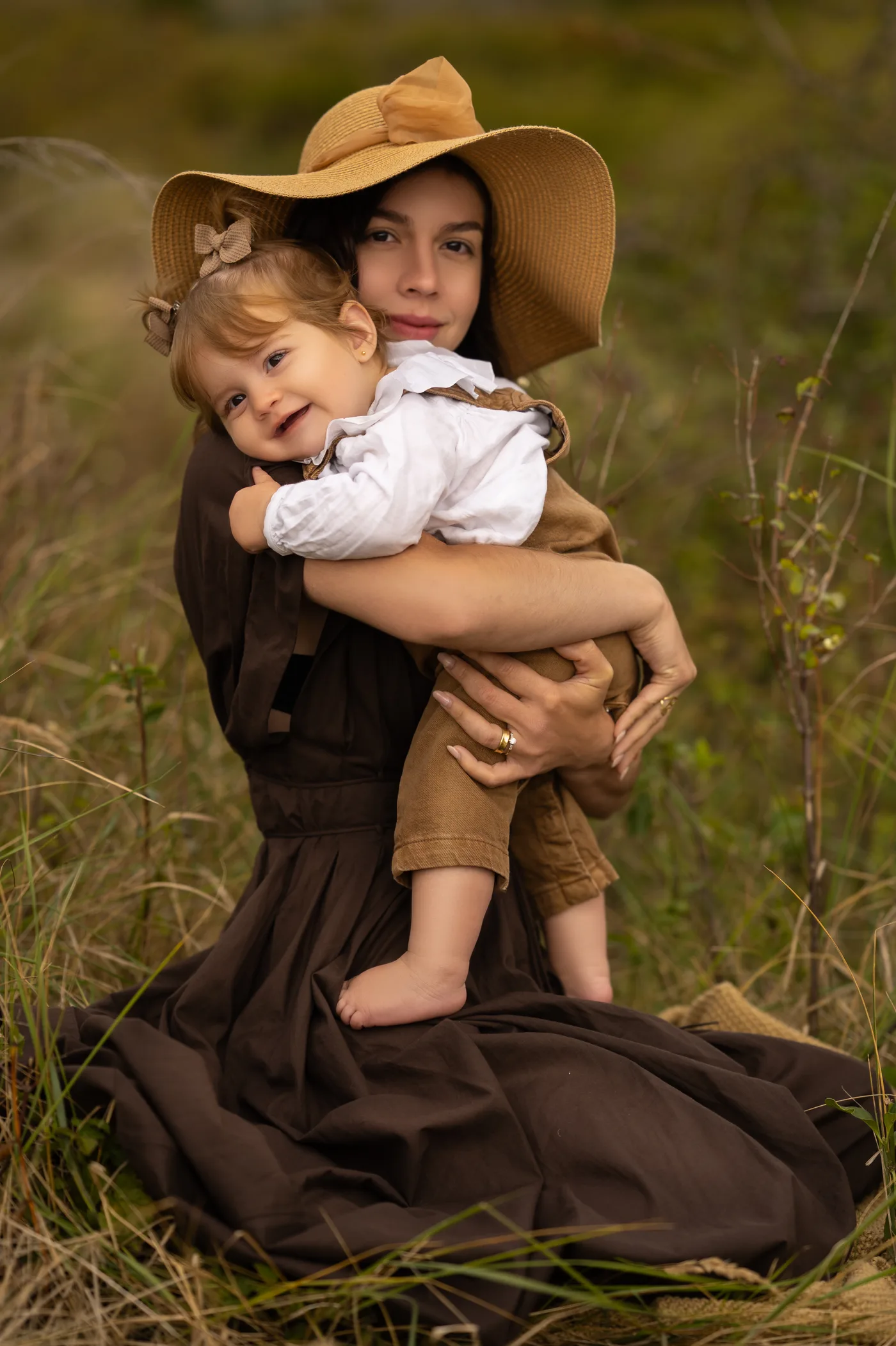 Mãe segurando a filha pequena no capim alto, as duas de chapéu