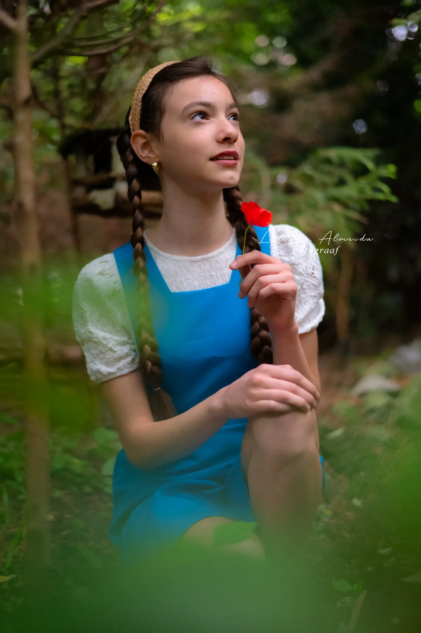 Menina de tranças e avental azul segurando uma flor vermelha na mata
