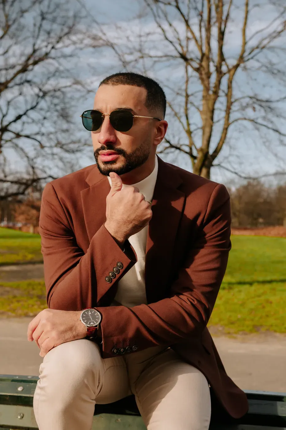 Retrato masculino de blazer terracota, mão junto ao rosto, no parque